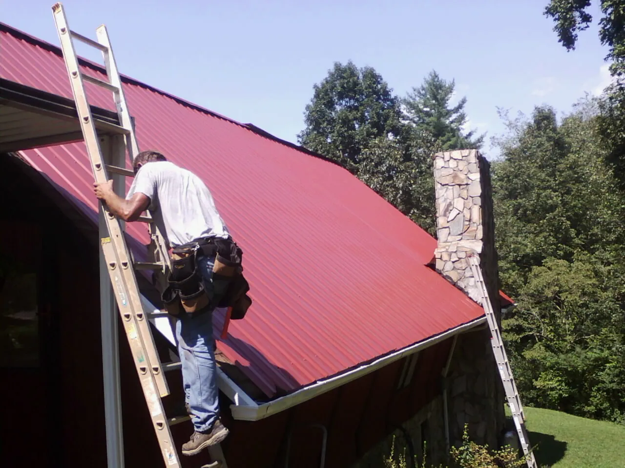 Roofer on a ladder beside a red metal roof and stone chimney — Soffit & Fascia Repair by Roofing Brothers in Scribner